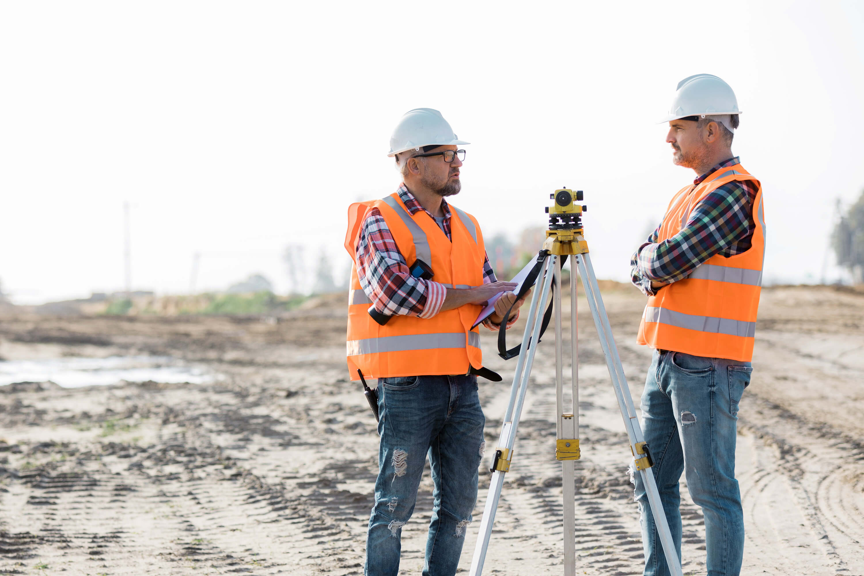 Two engineers wearing orange vests, using surveying equipment at a construction site.