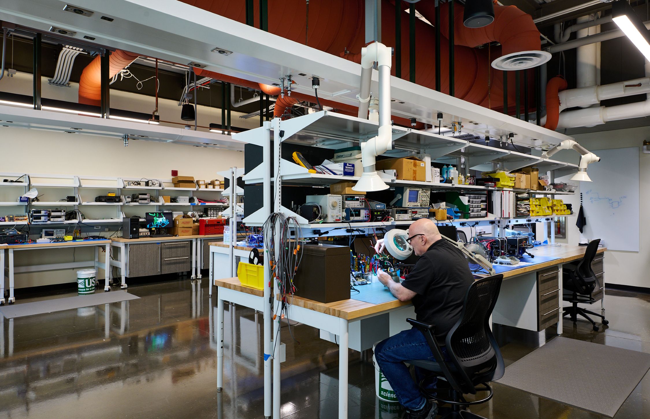 A technician works on electronic equipment at a laboratory workbench, surrounded by testing instruments and storage shelves