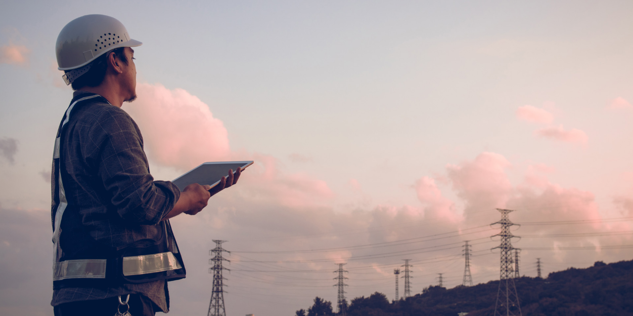 A construction engineer wearing safety gear, holding a tablet while inspecting an industrial site at sunset.